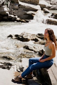 Traveler girl is sitting on the rock near waterfall and looking toward. Travelling in Karpathian mountains. Cascade waterfall. Beautiful landscape