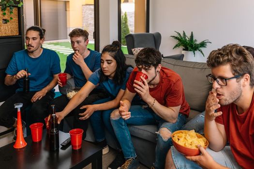 friends party at home watching television. group watching a movie or sporting event. young people eating and drinking. sitting on the sofa in the living room, natural light. table with drink and food.