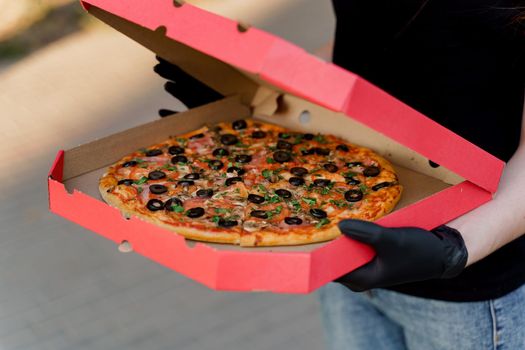 Pizza with salami and cheese in cardboard box. Safety delivery in black medical gloves from restaurant. Quarantine for stop coronavirus covid-19