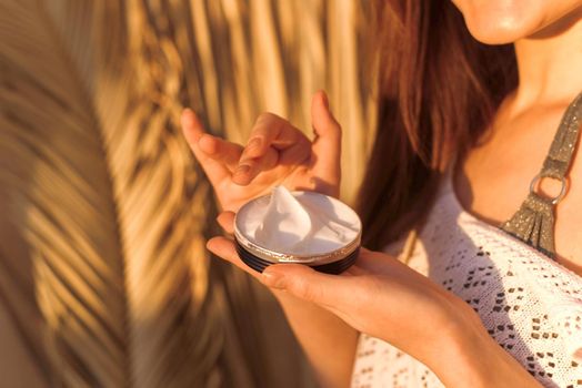 The hands of a young woman hold a moisturizing body cream. Sunlight, shadows of tropical palm leaves