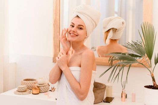 A young woman comes out of the shower wrapped in a towel, smiling and looking at the camera. Spa treatments at home, skin care