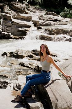 Traveler girl is sitting on the rock near waterfall and looking toward. Travelling in Karpathian mountains. Cascade waterfall. Beautiful landscape