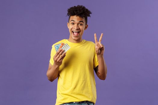 Health, influenza, covid-19 concept. Portrait of upbeat, healthy young man feel much better after taking vitamins or tablets, hold prescribed drugs, show peace sign, smiling pleased.