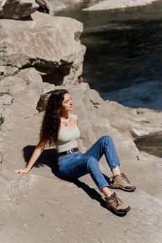 Traveler girl is sitting on the rock near waterfall and looking toward. Travelling in Karpathian mountains. Cascade waterfall. Beautiful landscape