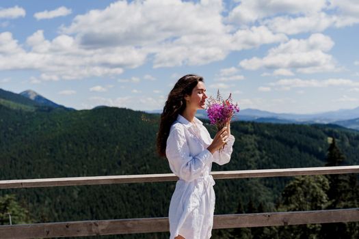 Attractive girl with bouquet of flowers on the green hills background. Young woman weared white dress is holding pink flowers