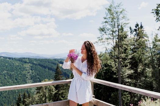 Attractive girl with bouquet of flowers on the green hills background. Young woman weared white dress is holding pink flowers