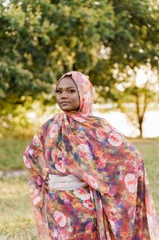 Muslim woman African ethnicity weared traditional colorful hijab on the green meadow. Pretty black girl smiles.