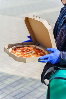 Pizza with salami and cheese in cardboard box. Safety delivery in blue medical gloves from restaurant. Quarantine for stop coronavirus covid 19