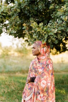 Muslim black woman African ethnicity weared traditional colorful hijab smiles and looks left side under green tree. Pretty black girl smiles.