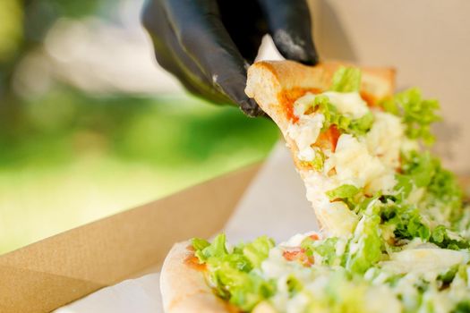 Pizza close-up in hands in medical gloves. Courier in medical mask holds pizza in hands and looks to pizza with green salad, tomatoes, cheese