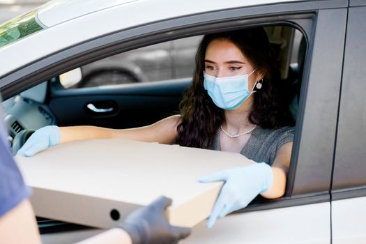 Girl in medical mask and gloves gets huge pizza in car. Big size pizza in carton eco box. Courier gives large pizza to young business woman. Delivery by car