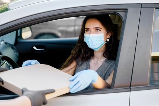 Beautiful young girl seats in car and gets pizza from courier. Woman in medical mask and gloves. Italian hot food delivery. Delivering by car.