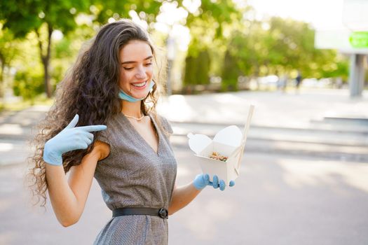 Young surprised girl with curly hair in medical gloves and mask holds wok in box in hands and smiles. Udon noodles in white box delivery.