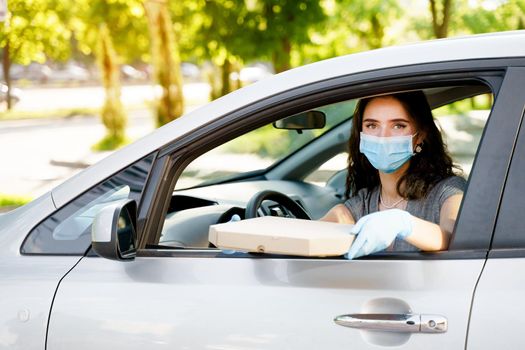 Safe food delivery from pizzeria to car during quarantine coronavirus. Attractive business woman in medical mask and gloves gets pizza in cardboard box