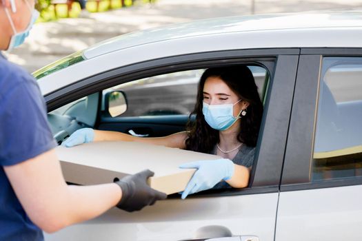Girl in medical mask and gloves gets huge pizza in car. Big size pizza in carton eco box. Courier gives large pizza to young business woman. Delivery by car
