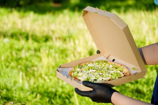 Pizza close-up in hands in medical gloves. Courier in medical mask holds pizza in hands and looks to pizza with green salad, tomatoes, cheese