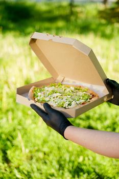 Pizza close-up in hands in medical gloves. Courier in medical mask holds pizza in hands and looks to pizza with green salad, tomatoes, cheese