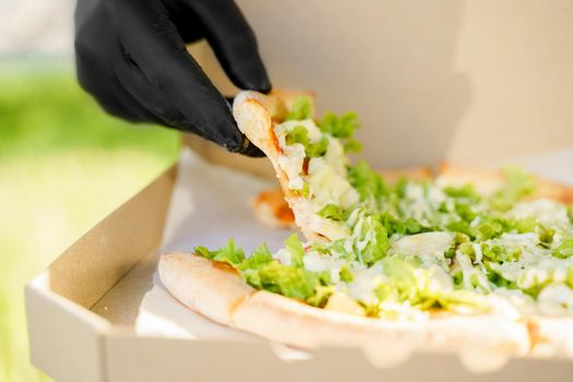 Pizza close-up in hands in medical gloves. Courier in medical mask holds pizza in hands and looks to pizza with green salad, tomatoes, cheese