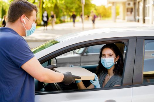 Safe food delivery from pizzeria to car during quarantine coronavirus. Attractive business woman in medical mask and gloves gets pizza in cardboard box