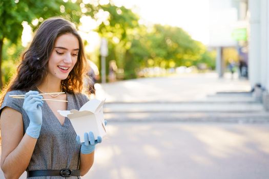 Young surprised girl with curly hair in medical gloves and mask holds wok in box udon noodles in hands and smiles. Udon noodles in white box delivery.