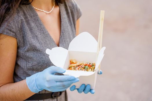 Closeup tasty food wok in box delivery. Girl holds udon noodles with in hands in medical gloves. Japanese spicy food in disposable eco boxes.