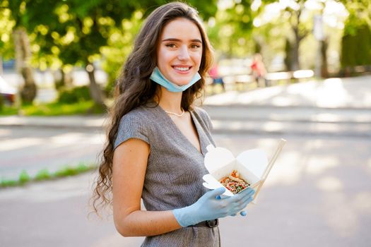 Young chinese girl with curly hair in medical gloves and mask holds wok in box in hands and smiles. Udon noodles in white box delivery