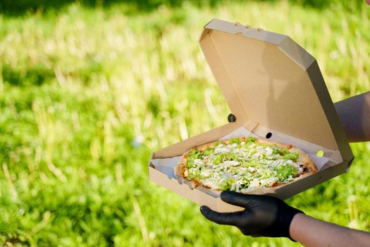 Pizza close-up in hands in medical gloves. Courier in medical mask holds pizza in hands and looks to pizza with green salad, tomatoes, cheese