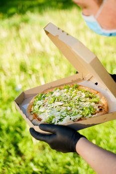 Pizza close-up in hands in medical gloves. Courier in medical mask holds pizza in hands and looks to pizza with green salad, tomatoes, cheese