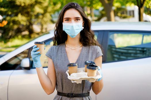 Girl in medical mask holds 4 coffee in hands with gloves. One cup of coffee in hands. Delivery by car. Safe delivery.