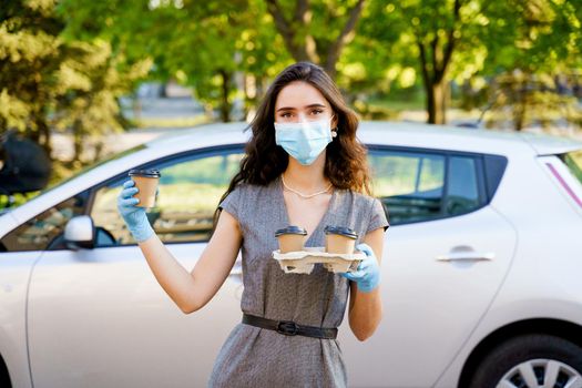 Girl in medical mask holds 4 coffee in hands with gloves. One cup of coffee in hands. Delivery by car. Safe delivery.