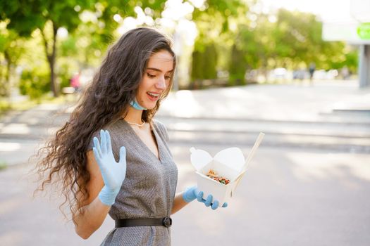 Udon noodles in white box delivery. Young surprised girl in gloves and mask holds wok in box in hands and smiles.