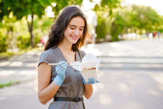 Young surprised girl with curly hair in medical gloves and mask holds wok in box in hands and smiles. Udon noodles in white box delivery