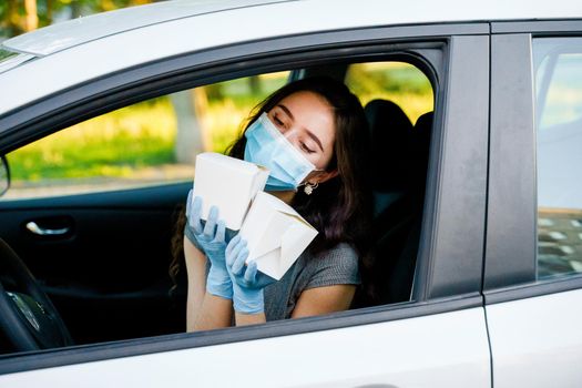 Young attractive girl in car in medical gloves and mask holds wok in box udon noodles in hands and smiles. Udon noodles in white box delivery