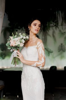 Bride with wedding bouquet smiles and touches her face. Attractive girl portrait for social networks. Girl in wedding dress in luxury restaurant.