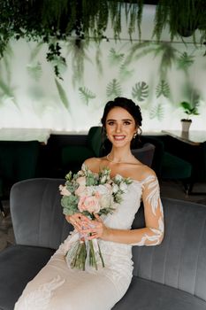 Bride with wedding bouquet smiles, looks in camera and touches her face. Attractive girl portrait. Girl in wedding dress in luxury restaurant