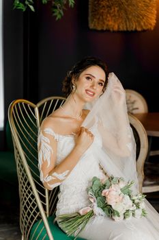 Bride with wedding bouquet smiles, looks in camera and touches her face. Attractive girl portrait. Girl in wedding dress in luxury restaurant