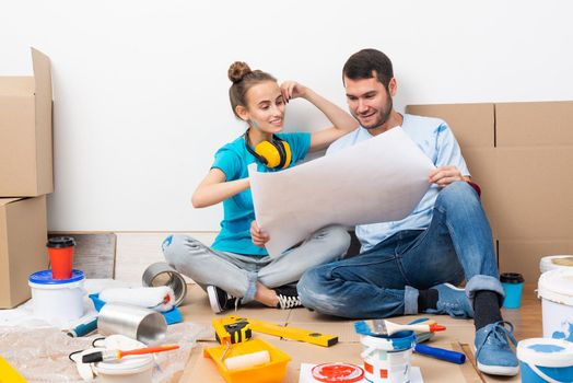 Young man and woman together planning their home renovation. Cardboard boxes, painting tools and materials on floor. House remodeling and interior renovation. People looking at blueprint at home.