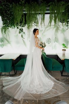 Young attractive bride in wedding dress with bridal veil looks at bouquet and smiles. Girl in wedding gown in modern restaurant