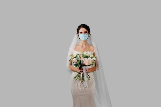 Bride in medical mask and wedding bouquet at coronavirus covid-19 quarantine period. Attractive girl in studio