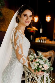 Bride with wedding bouquet smiles, looks in camera and touches her face. Attractive girl portrait for social networks. Girl in wedding dress in luxury restaurant