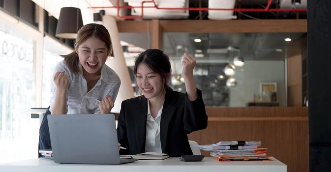 Two young Asian businesswomen show joyful expression of success at work smiling happily with a laptop computer in a modern office..