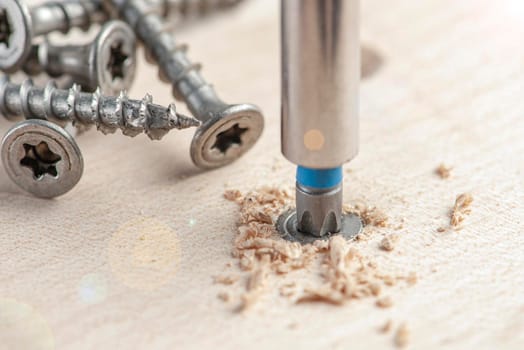 Screw the screw into the board. Furniture production, a screwdriver twists a self-tapping screw into a board. Several silver screws lie on the desktop.