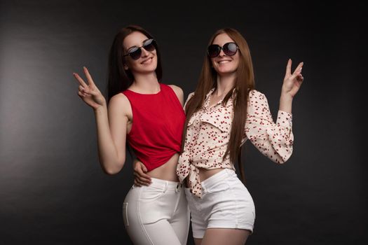 Two happy sisters in sunglasses holding hands and jumping. Brunette models in casual clothes with open belly getting fun together. Beautiful young friends in stylish looks and gesturing by fingers.