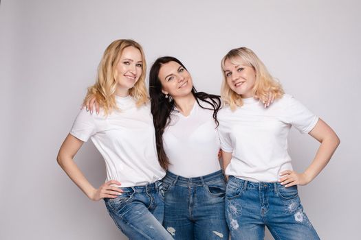 Side view of three beautiful young women standing one by one. Blonde and brunette girls in white t-shirts and jeans posing at camera. Happy friends in casual clothes smiling being close to each other.
