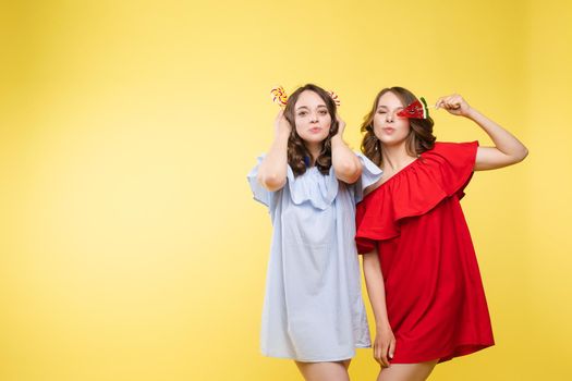 Front view of charming twins in pink dresses closing eyes with lollipops and posing on isolated background. Two female friends laughing and having fun in studio. Concept of childhood and beauty.