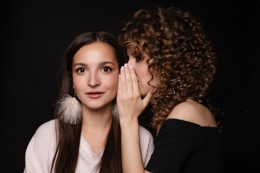 Front view of shocked brunette with big eyes and open mouth in white dress looking at camera while female friend whispering secret on black isolated background. Young women gossiping in studio.