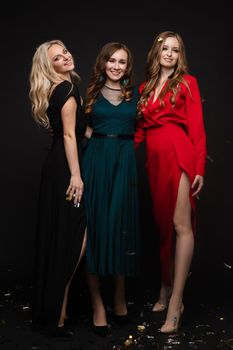 Studio full length portrait of three stunning young ladies in evening gowns posing for camera. Black background. New Year evening celebration birthday concept.