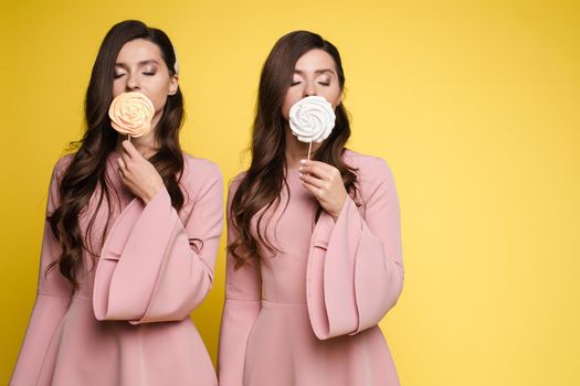 Front view of charming twins in pink dresses closing eyes with lollipops and posing on isolated background. Two female friends laughing and having fun in studio. Concept of childhood and beauty.