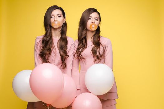 Young twins in elegant dresses holding pink and white balloons in their hands. Brunette sisters with long hair blowing big bubbles from chewing gum. Beautiful girls posing at camera with decorations.