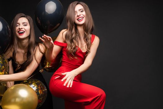 Happy young girls laughing and getting fun together. Brunette beautiful sisters in elegant clothes celebrating birthday. Gorgeous women with red lips standing near bunch of gold and black balloons.
